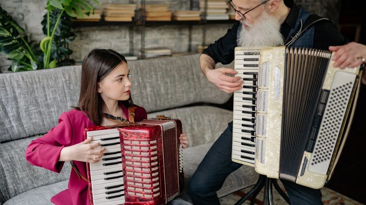 professor de barba branca e aluna tocando acordeon sentados em um sofá cinza durante a aula