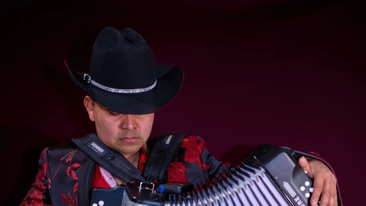 músico de chapéu preto e jaqueta vermelha tocando um acordeon de botões no palco