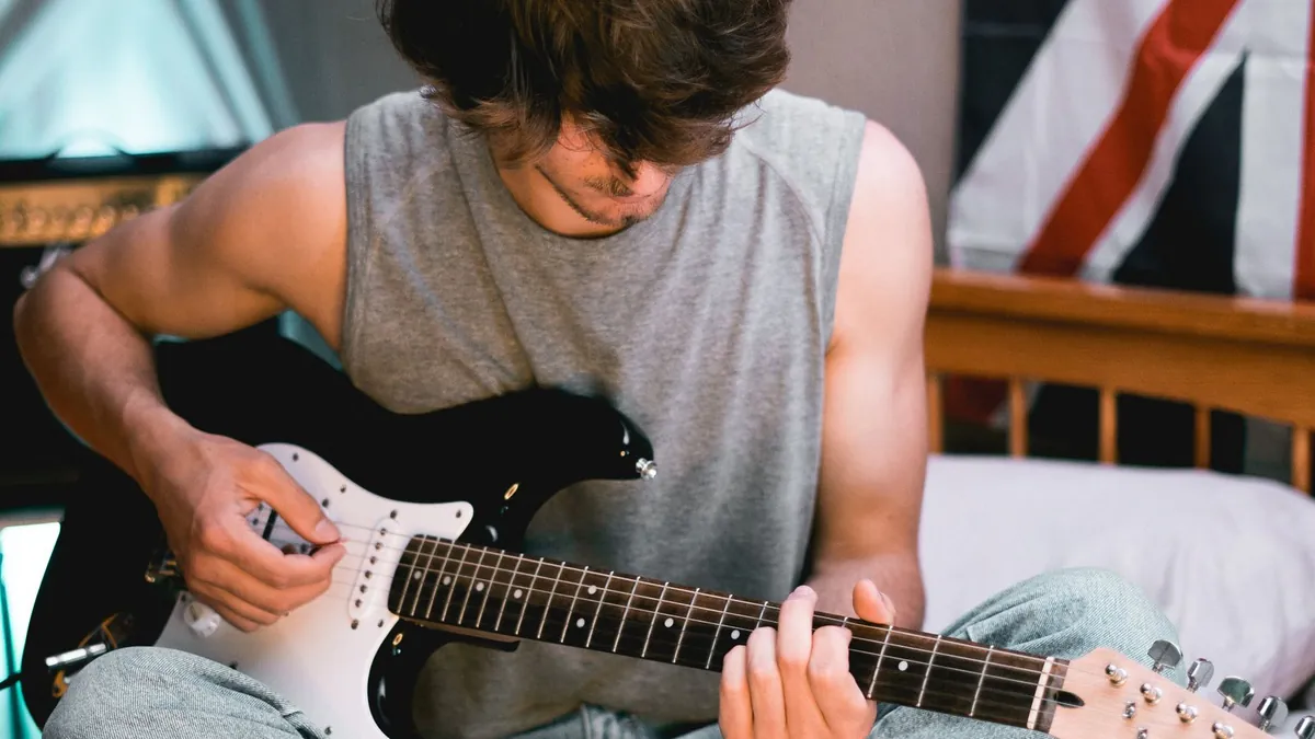 rapaz tocando guitarra elétrica branca e preta sentado em um quarto com bandeira britânica ao fundo
