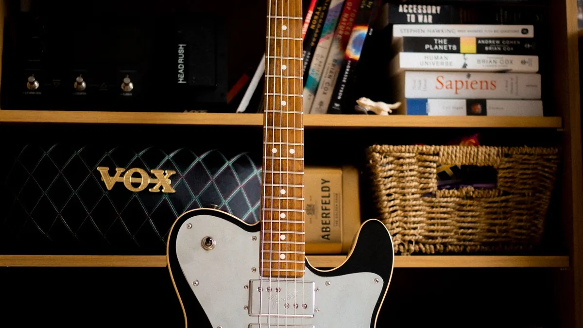 guitarra Telecaster branca e preta em pé diante de uma estante com livros e um amplificador Vox