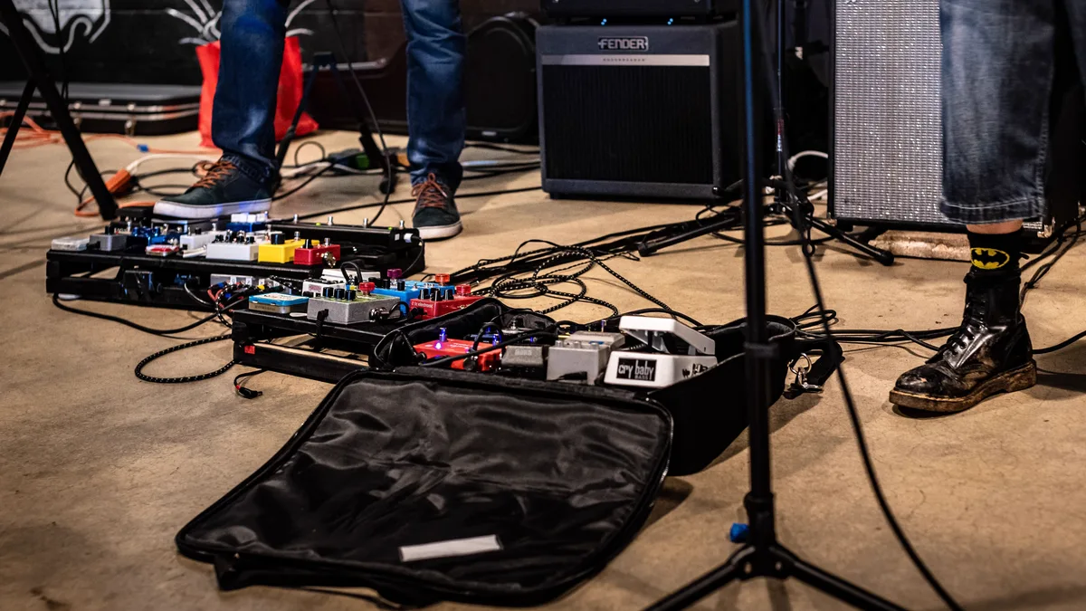 pedalboard no chão de um palco pequeno, com músico em pé ao lado de caixas e microfone
