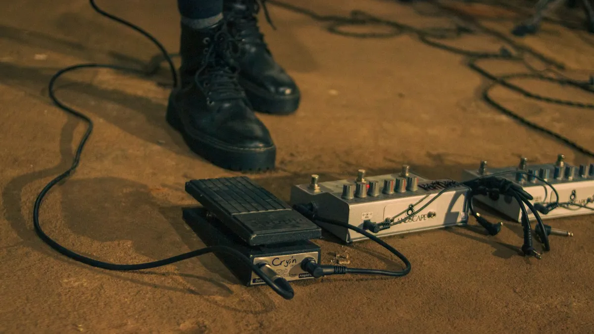 pedal de volume e pedaleira no chão de um palco com vários cabos soltos ao lado das botas de um guitarrista