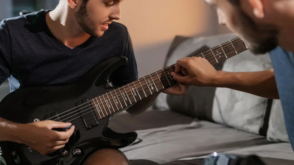 professor mostrando um acorde no braço da guitarra para um aluno sentado no sofá