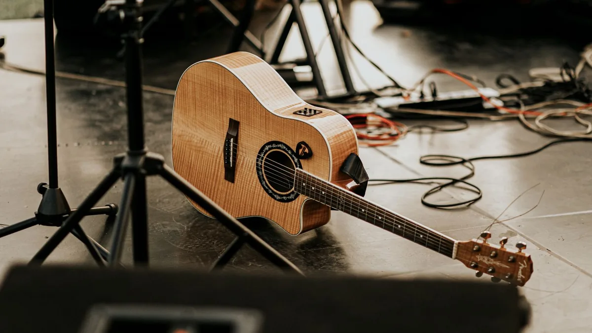 violão eletroacústico cutaway de madeira clara no chão de um palco, entre cabos e suportes de microfone