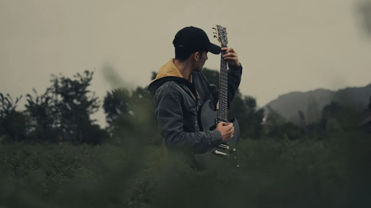 músico de boné e jaqueta segurando uma guitarra em um campo aberto ao entardecer