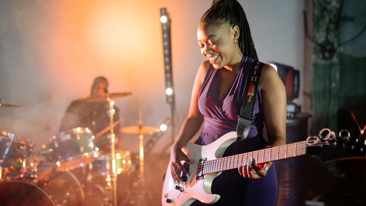 guitarrista tocando em pé no palco com uma guitarra branca presa por correia preta no ombro