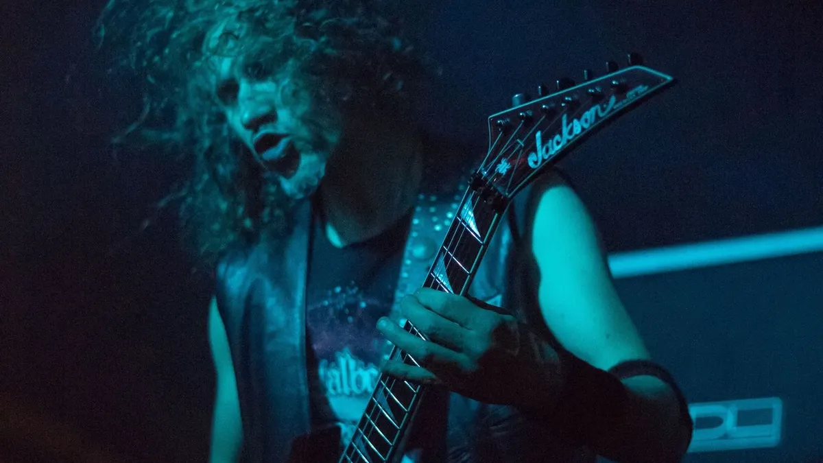 guitarrista com cabelo cacheado tocando uma guitarra pontuda em palco com luz azul