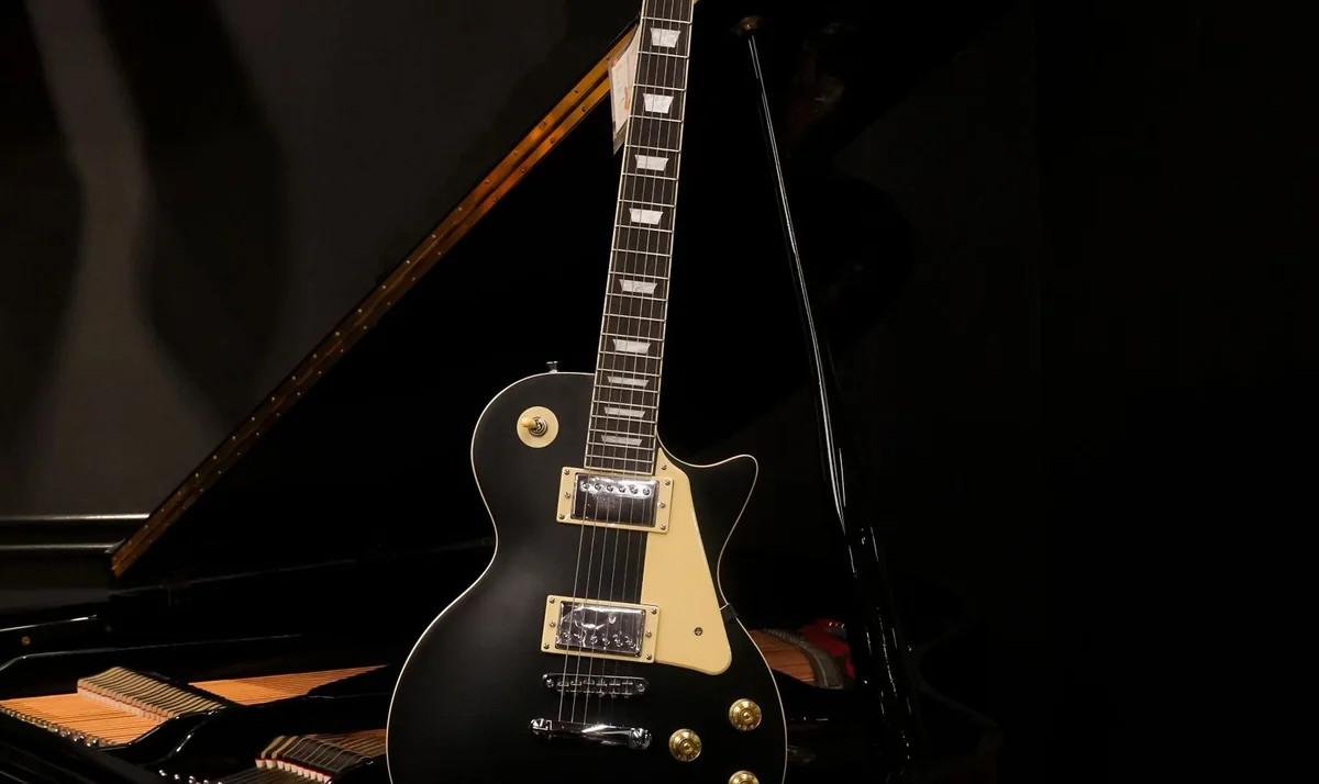 guitarra elétrica preta, modelo Les Paul, apoiada em um piano de cauda sob luz baixa