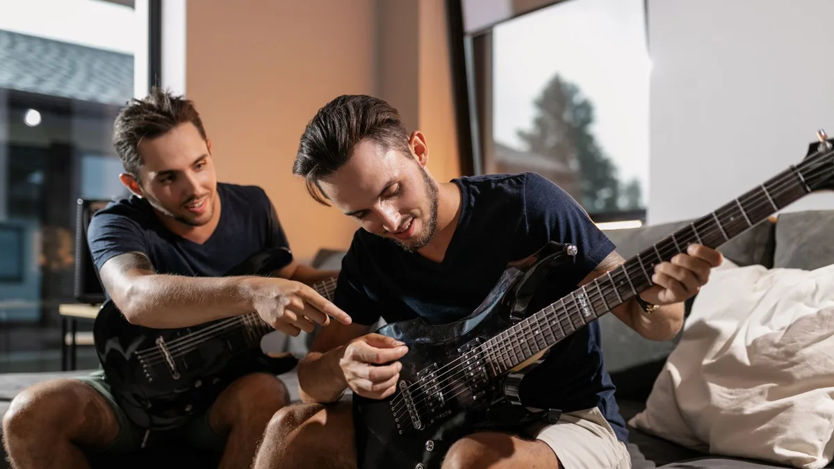 dois amigos sentados em um sofá, um deles tocando uma guitarra preta enquanto o outro aponta para o braço