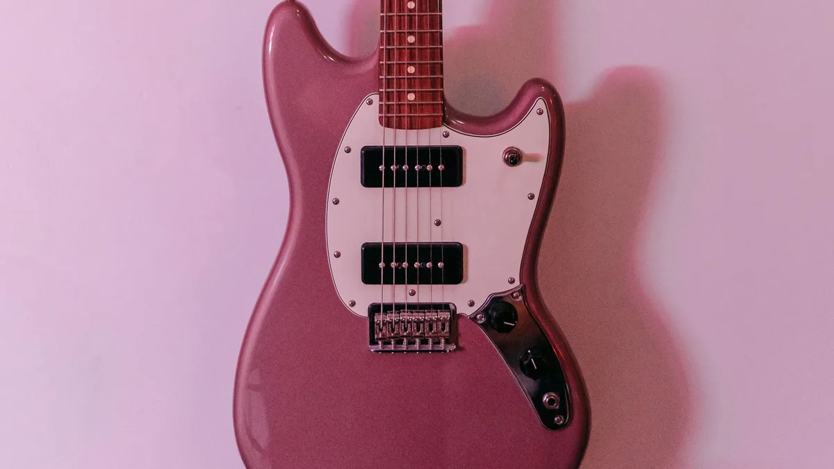 guitarra rosa metálico estilo Mustang pendurada em parede lilás com dois captadores e escudo branco