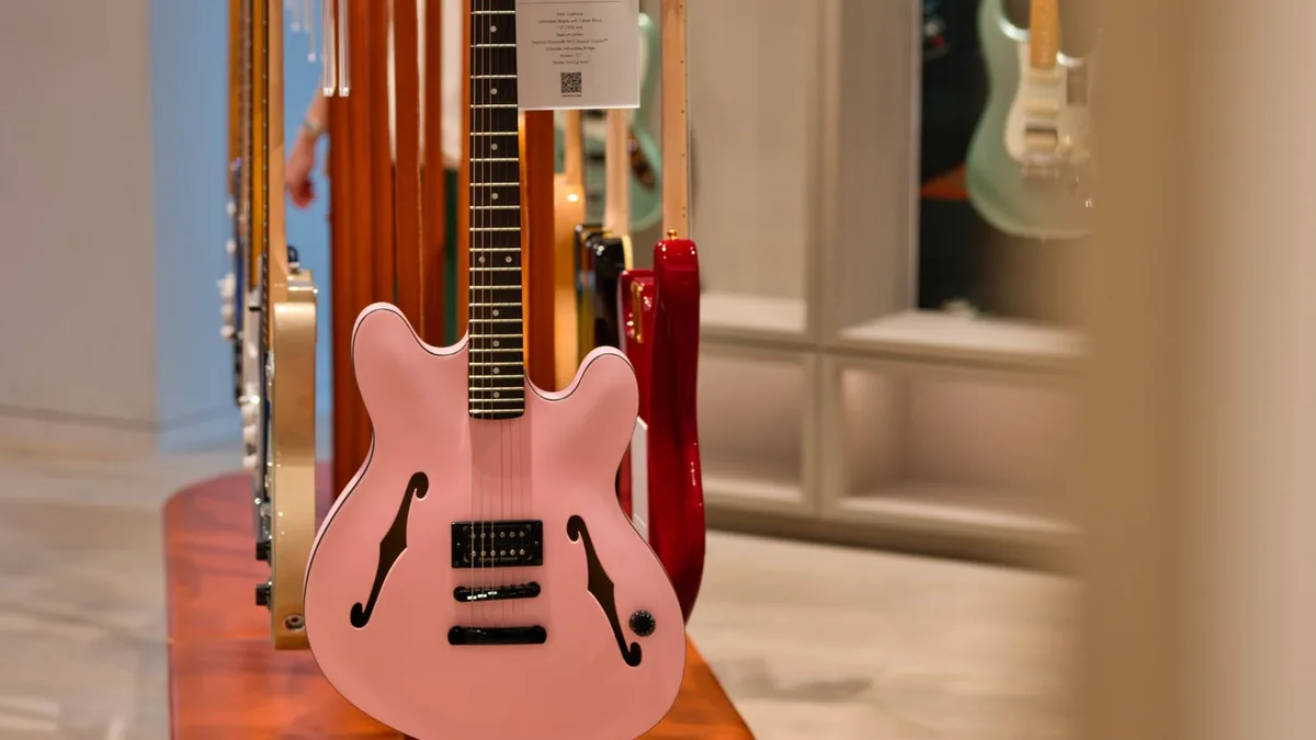guitarra semi-acústica rosa clara exposta em suporte de madeira em loja de instrumentos, com outras guitarras ao fundo