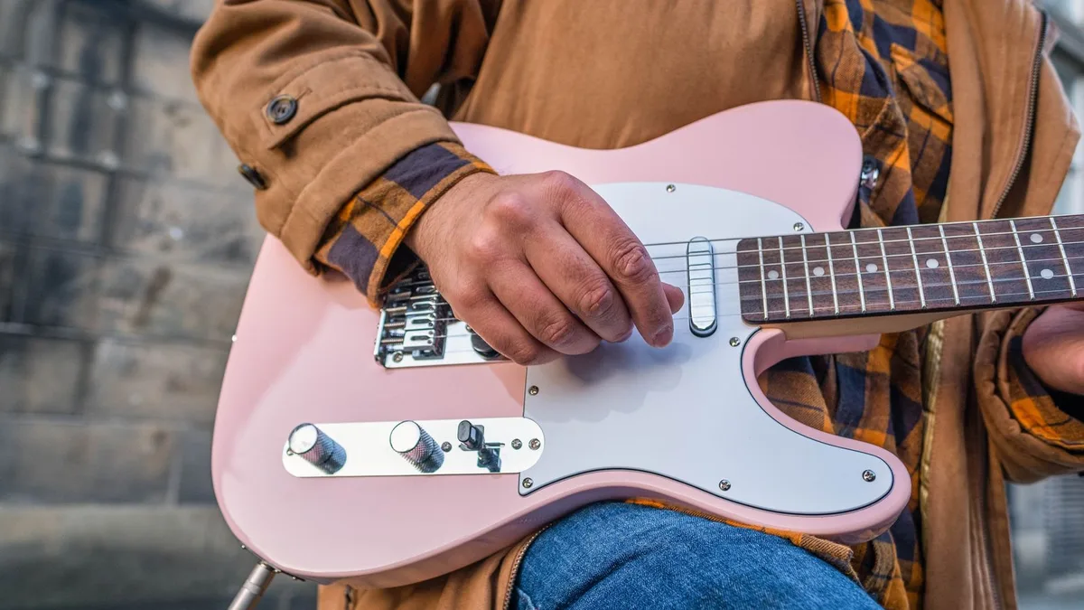 músico de jaqueta tocando uma guitarra rosa pastel estilo Telecaster ao ar livre