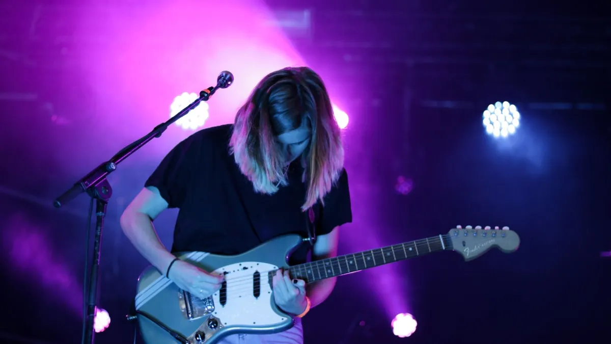 guitarrista de cabelo comprido tocando uma guitarra azul clara sob luz roxa de palco