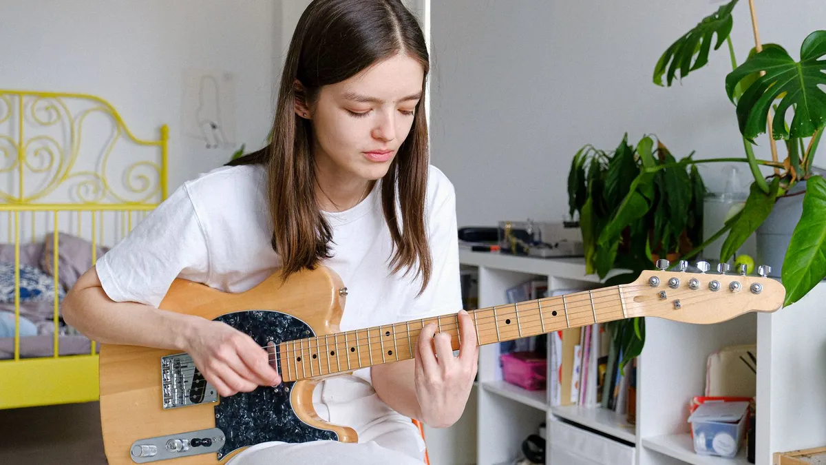 jovem de camiseta branca tocando uma guitarra de corpo claro em um quarto com estante e plantas