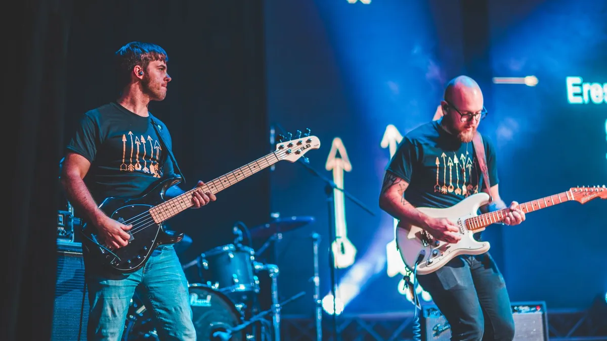 dois guitarristas tocando em um palco com iluminação azul, um com guitarra escura e outro com guitarra clara
