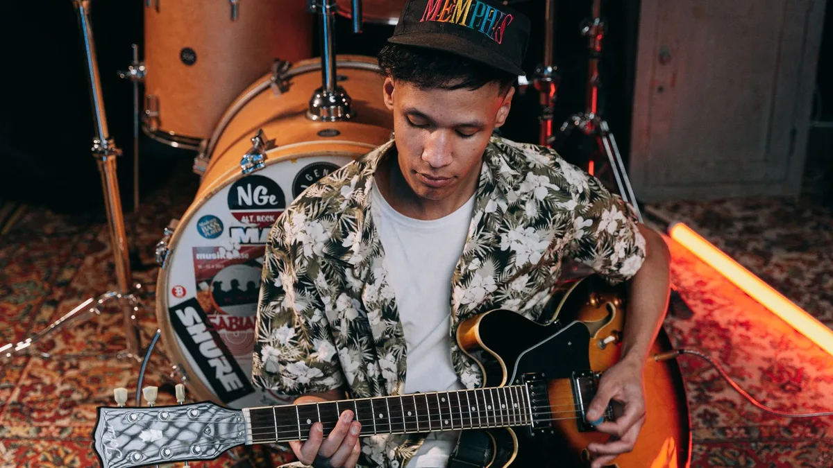 músico de boné e camisa estampada tocando uma guitarra em uma sala com bateria e luz de neon