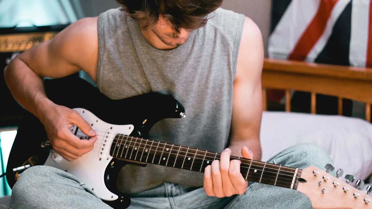 rapaz sentado na cama tocando uma guitarra preta com escudo branco, com um caderno ao lado