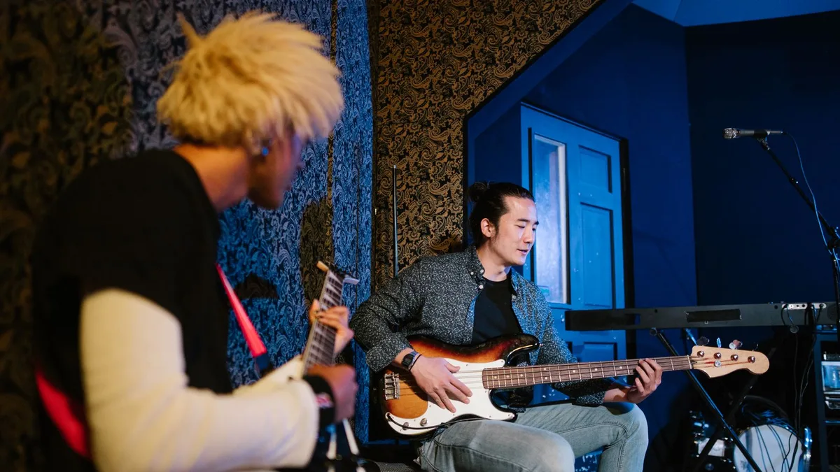 dois músicos em um estúdio com paredes azuis, um deles de cabelo loiro e o outro sentado tocando uma guitarra