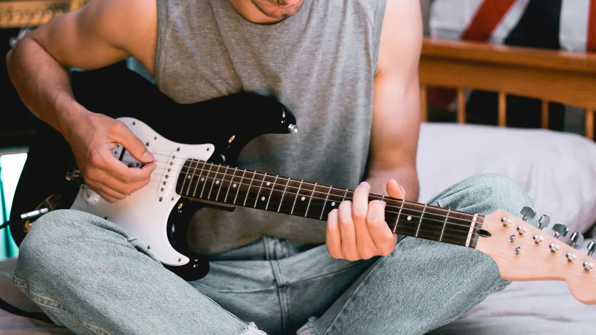 guitarrista sentado na cama tocando guitarra elétrica, com a mão esquerda no braço