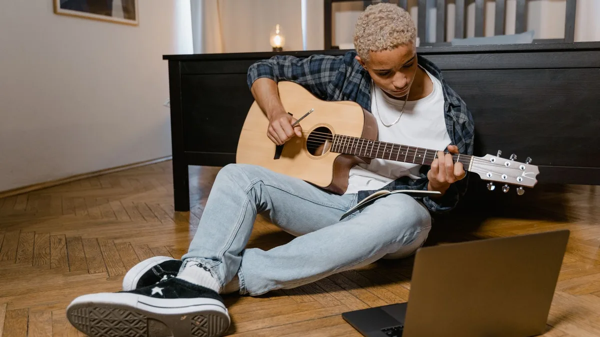 homem sentado no chão tocando violão com o notebook ao lado para aprender uma música