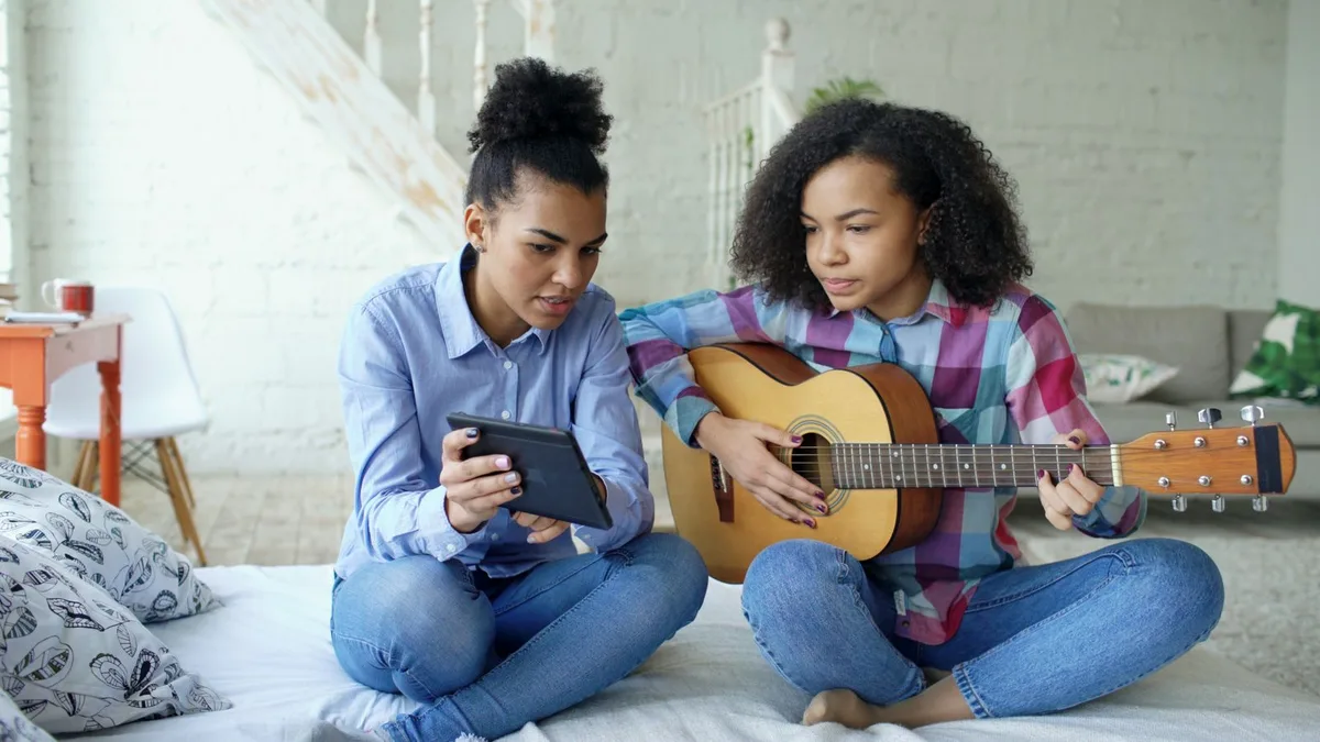 duas jovens sentadas na cama, uma com violão e outra lendo a cifra no tablet