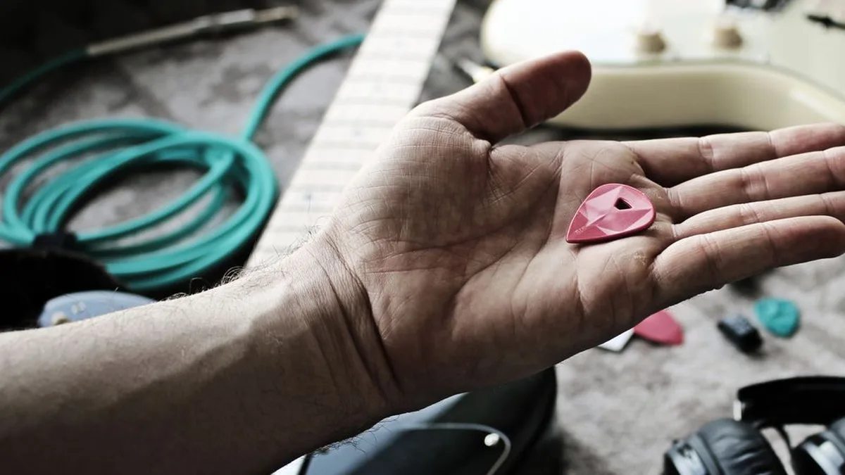 mão aberta segurando uma palheta rosa, com uma guitarra creme e um cabo verde ao fundo
