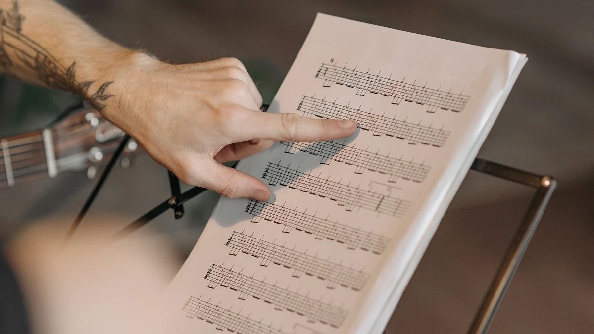 mão apontando para tablatura impressa em um livro, com o braço da guitarra ao lado