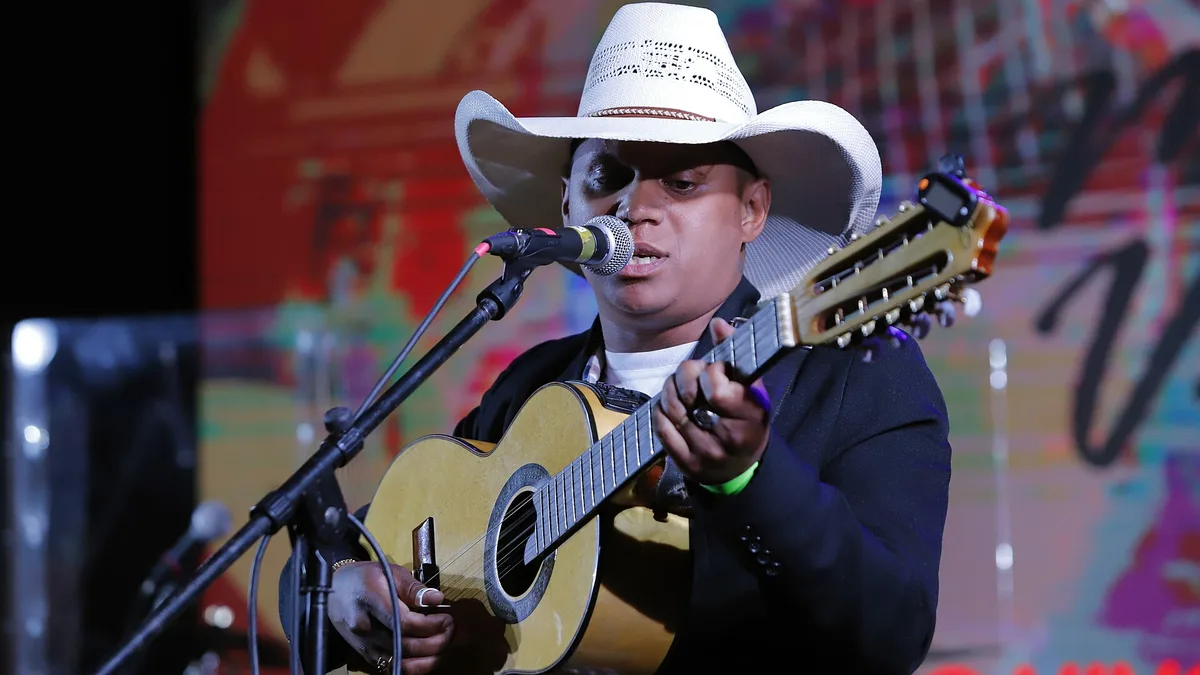 cantor de chapéu branco tocando uma viola caipira diante de um painel colorido em um festival