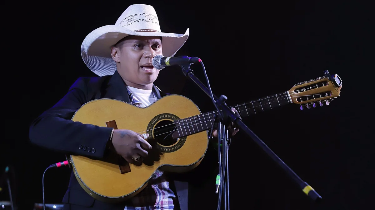 violeiro de chapéu branco cantando ao microfone e tocando uma viola caipira de dez cordas em um palco escuro