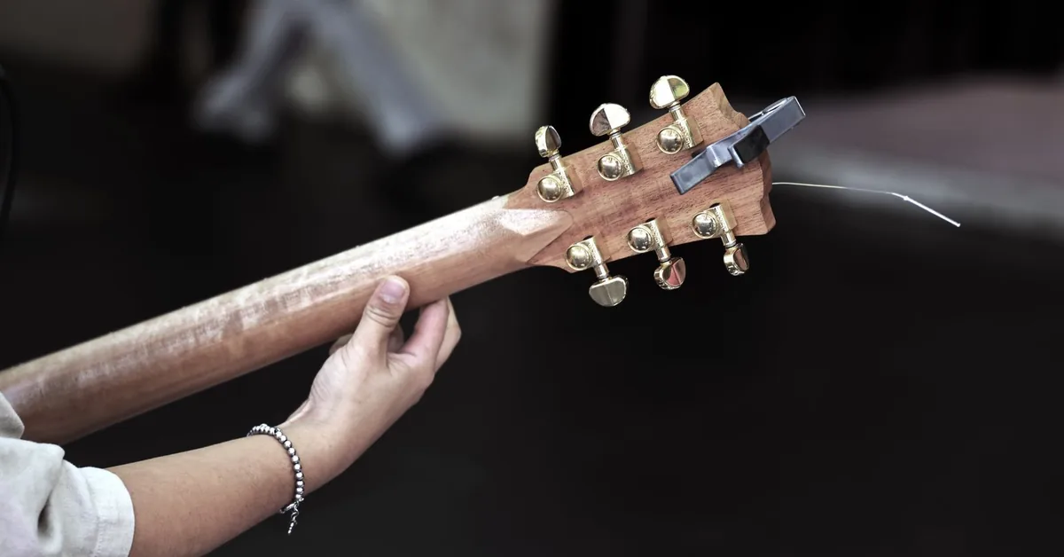 mãos segurando o braço de um violão em um palco escuro, com a cabeça e as tarraxas em evidência
