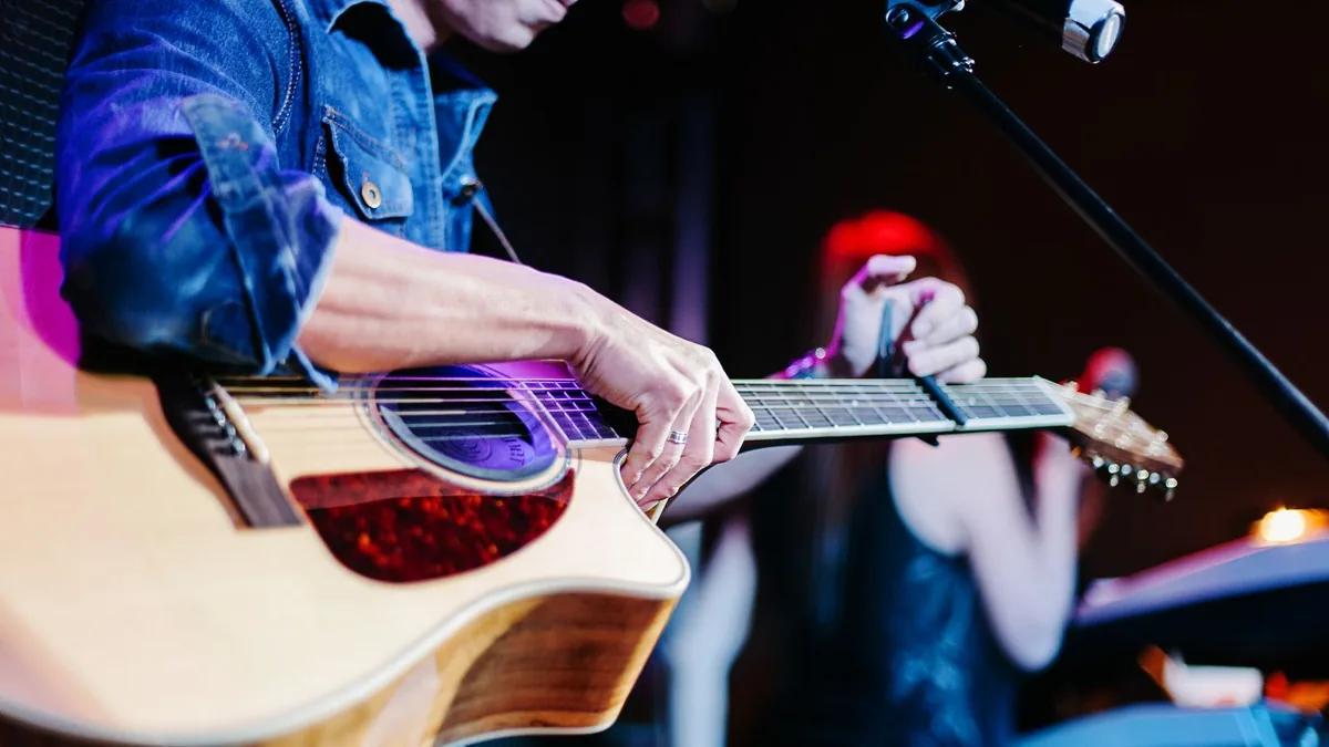 violonista de camisa jeans tocando um violão eletroacústico de corpo cutaway no palco, com microfone ao fundo