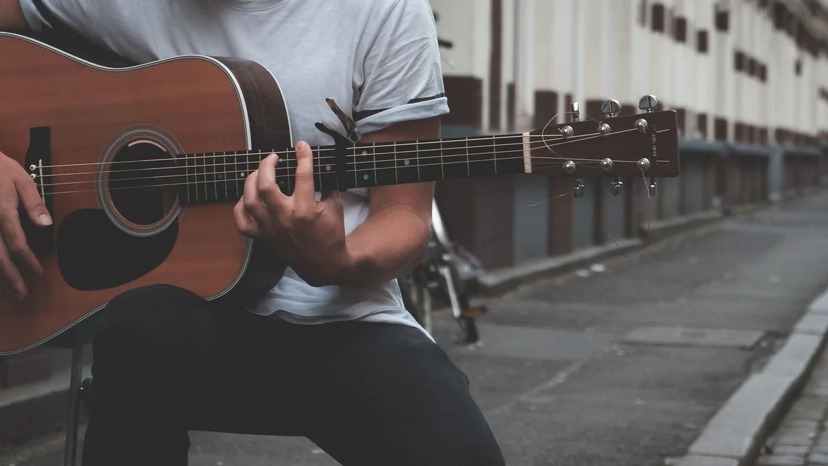 violonista de camiseta cinza tocando um violão de aço sentado em um banco, numa rua de cidade