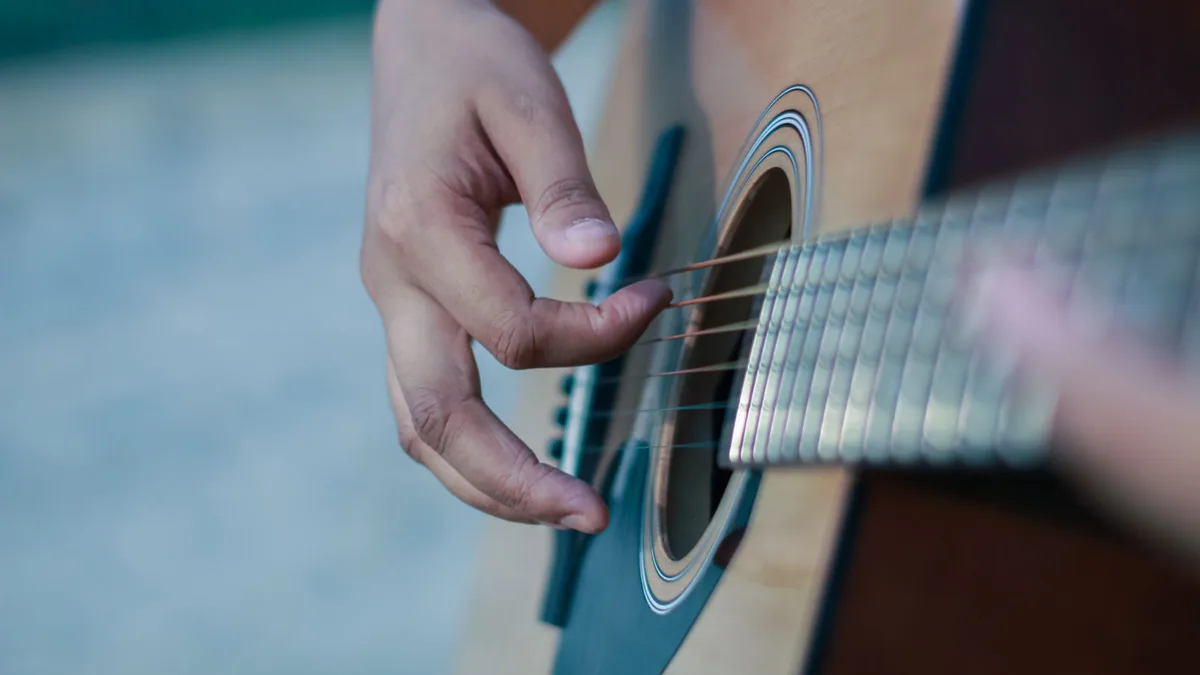 mão dedilhando as cordas de um violão de aço ao ar livre, com fundo desfocado