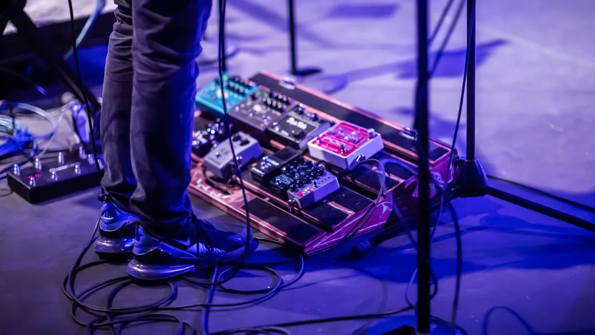 guitarrista em pé sobre uma pedalboard com vários pedais iluminada em tons de azul e roxo em um palco